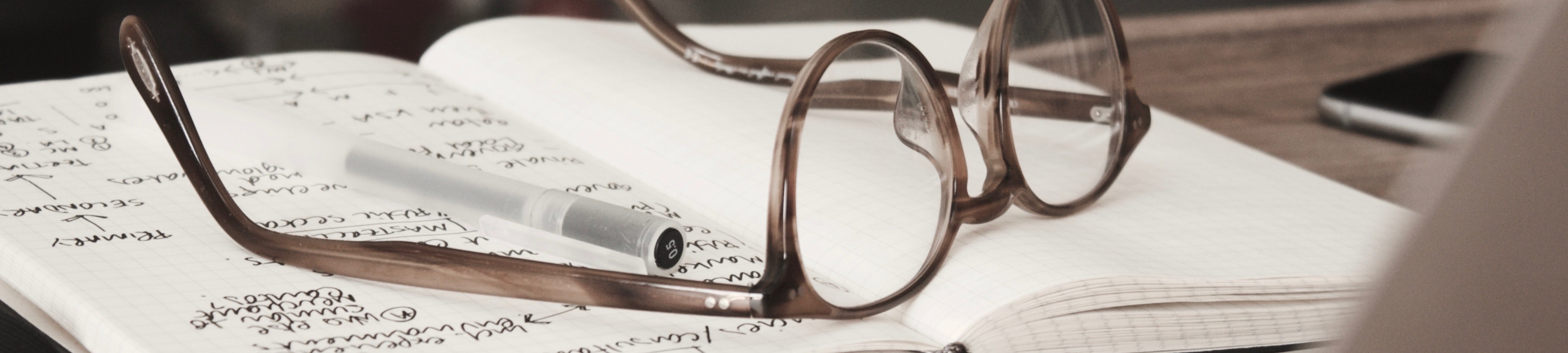 Photo of a pair of glasses and a pen resting on an open notebook. A phone and laptop are by the side on the desk