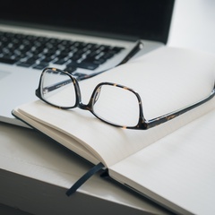 Pair of glasses and a pen lying on a notebook next to a laptop