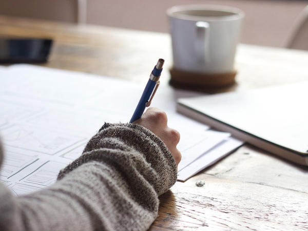A person writing in a book at a table with coffee and papers