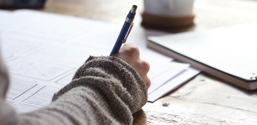 A person writing in a book at a table with coffee and papers