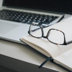 Pair of glasses and a pen lying on a notebook next to a laptop