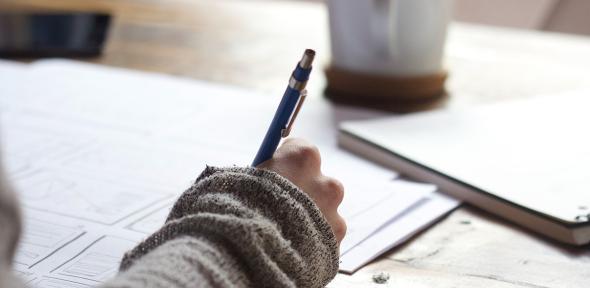 A person writing in a book at a table with coffee and papers