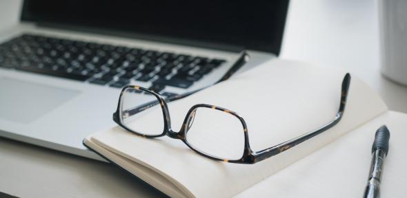 Pair of glasses and a pen lying on a notebook next to a laptop